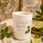 A white scented candle jar labeled 'Sweet Escape Luxury Candles Mint to Be' sits on a marble surface with mint leaves, lime slices, a beige cloth, and a framed quote in the background.